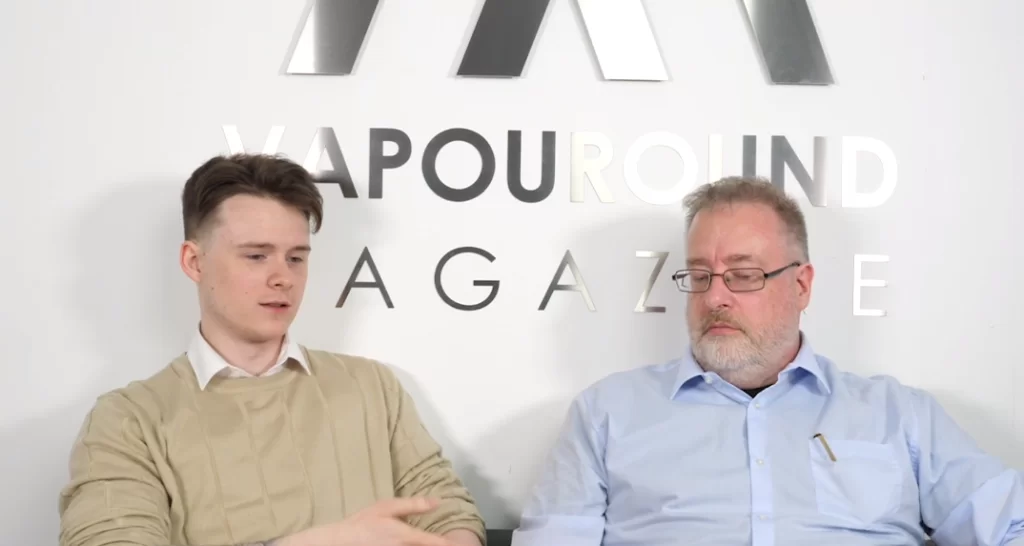 Two men sit beside each other. One is wearing a beige jumper and the other is in a blue shirt. They are in discussion in front of a sign that says Vapouround Magazine.