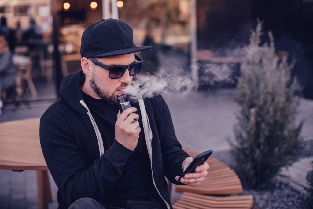 young man wearing a black hoodie and black cap sits at a table vaping