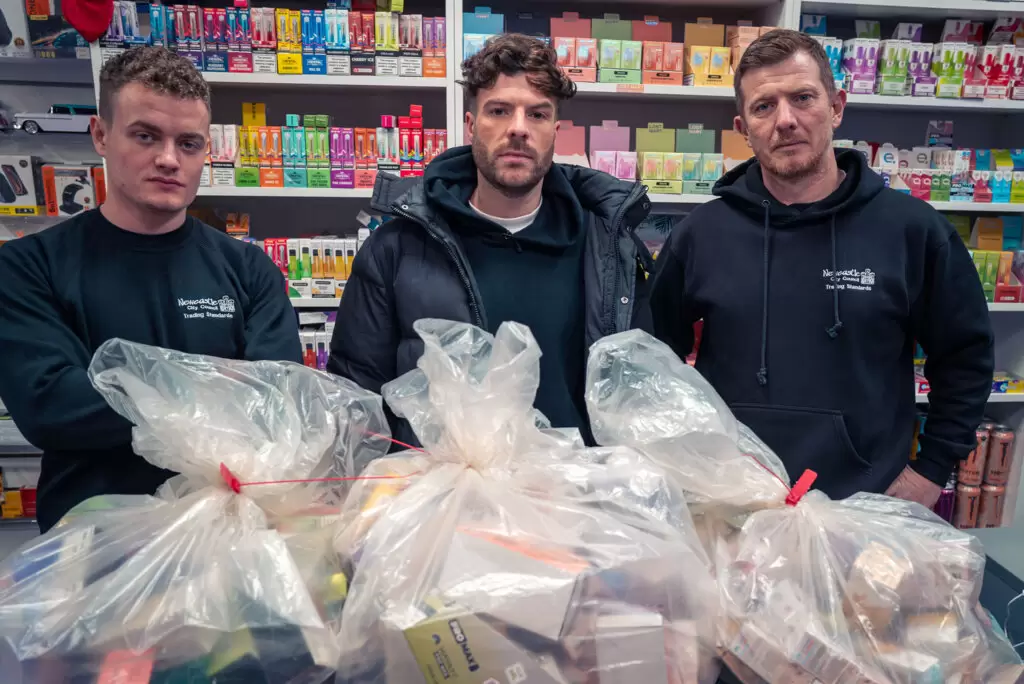 Jordan North with Connor and Paul from Newcastle Trading Standards in a corner shop surrounded by disposable vapes